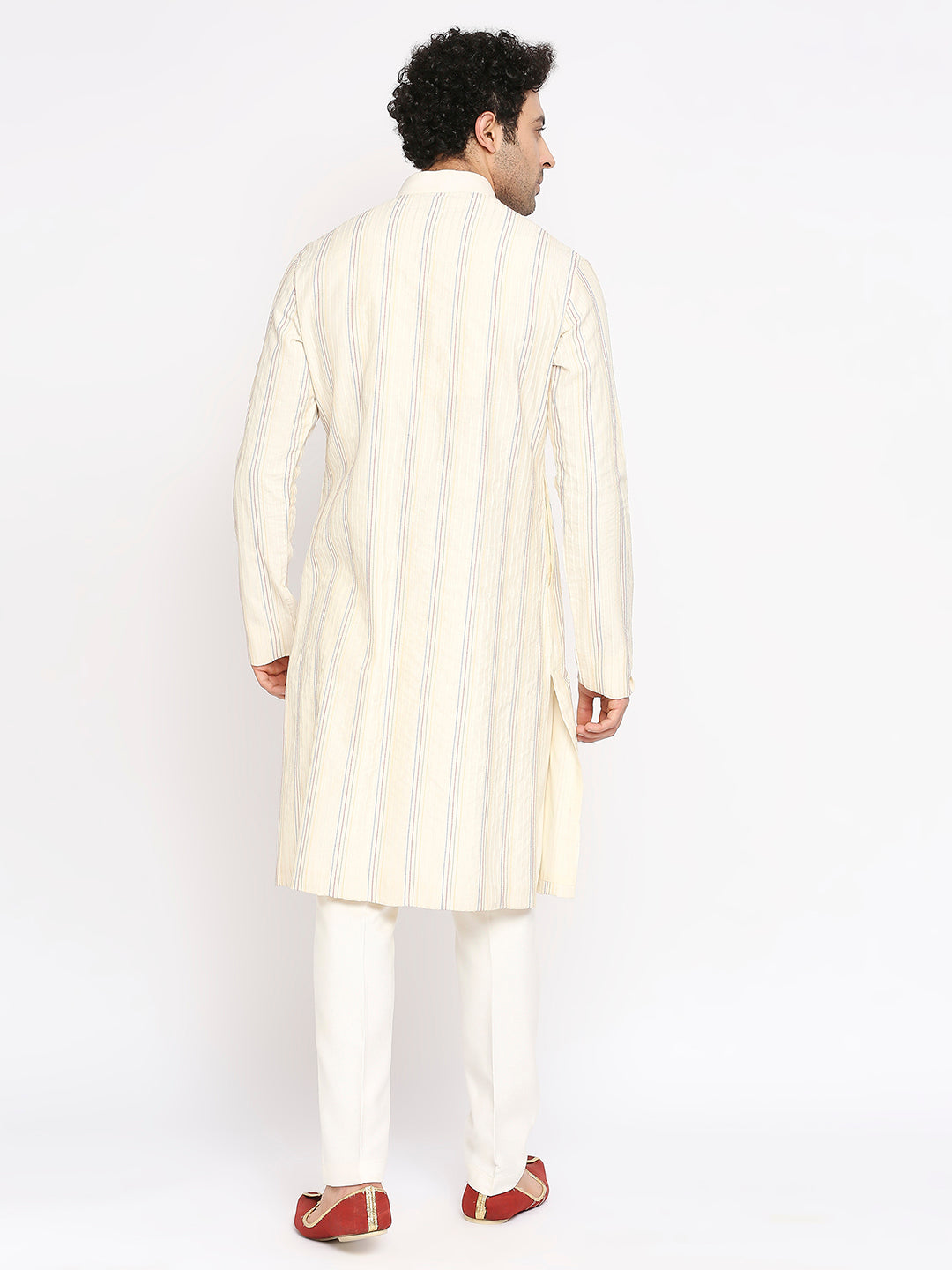 A man viewed from the back wearing a cream-colored kurta with thin vertical blue stripes, paired with white trousers and red shoes, against a plain white studio background.