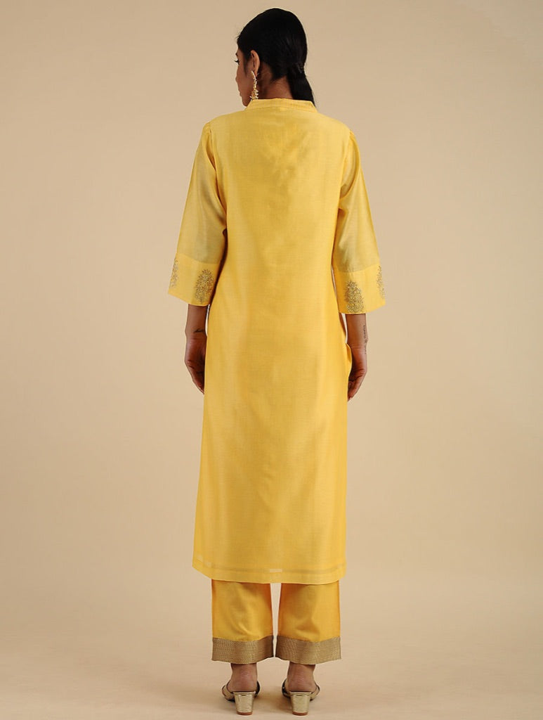 A woman viewed from the back wearing a bright yellow kurta and matching trousers with beige rolled hems, standing against a plain beige studio background.