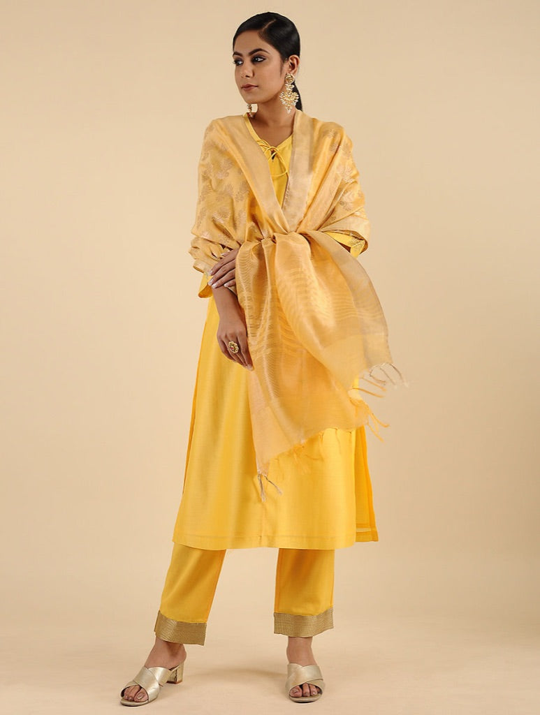 A woman models a traditional Indian outfit consisting of a yellow kurta, matching trousers, and a sheer yellow shawl, set against a solid beige studio background.