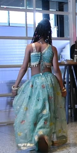 A woman viewed from behind wearing a light blue-green two-piece outfit with a floral print, embellished with gold sequins and fringe, in an indoor setting.