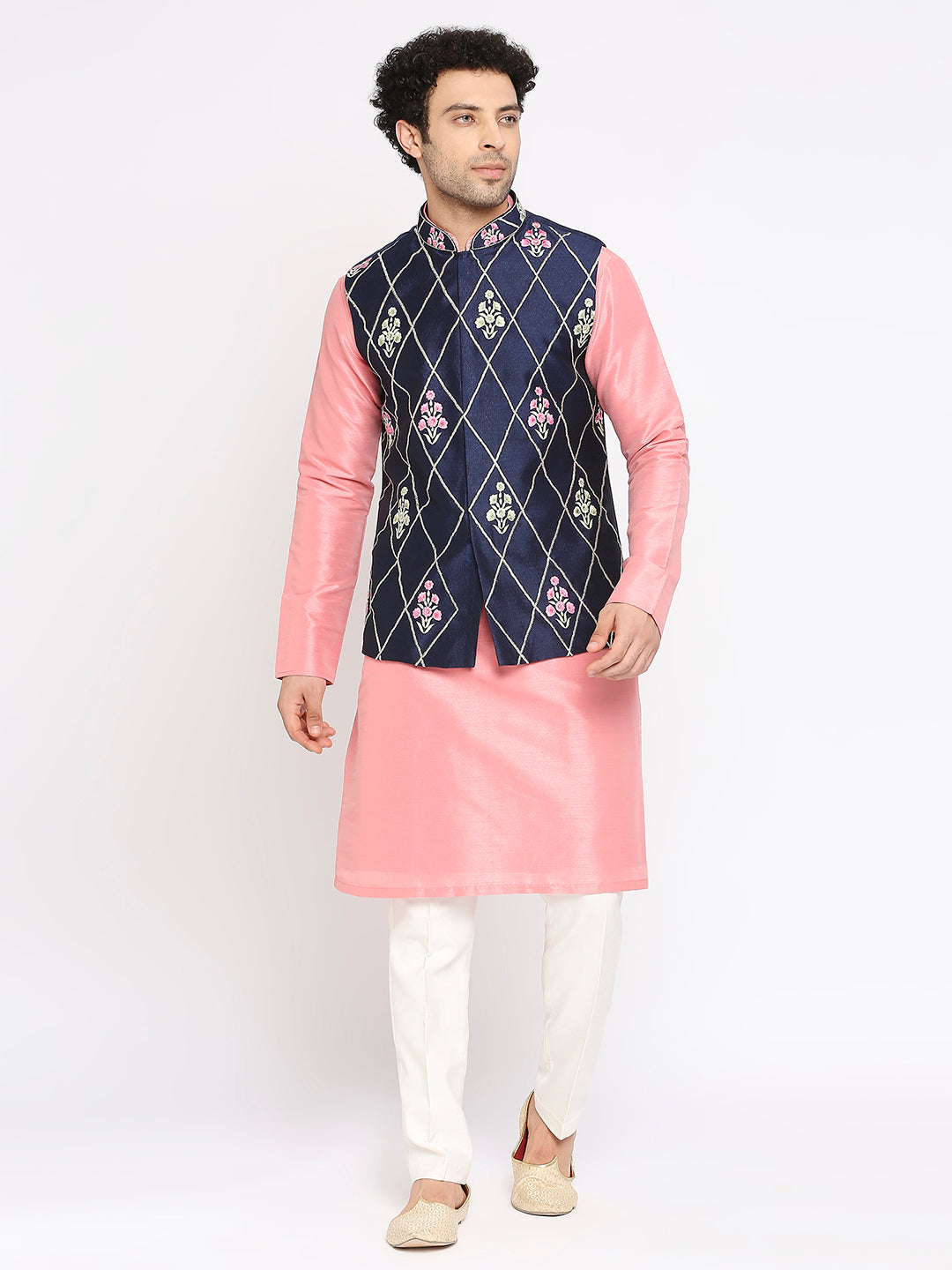 A man modeling a traditional Indian outfit consisting of a pink kurta, white trousers, and a navy blue vest with a diamond pattern of pink, white, and green floral embroidery, set against a plain white studio background.