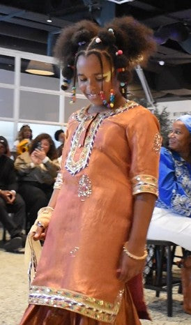 A woman modeling a traditional orange-brown embroidered tunic with metallic embellishments, standing in an indoor event space with seated spectators in the background.
