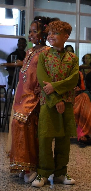 A young boy in a green traditional outfit with a floral embroidered vest and a woman in a peach and gold embroidered dress standing together in an indoor event space.