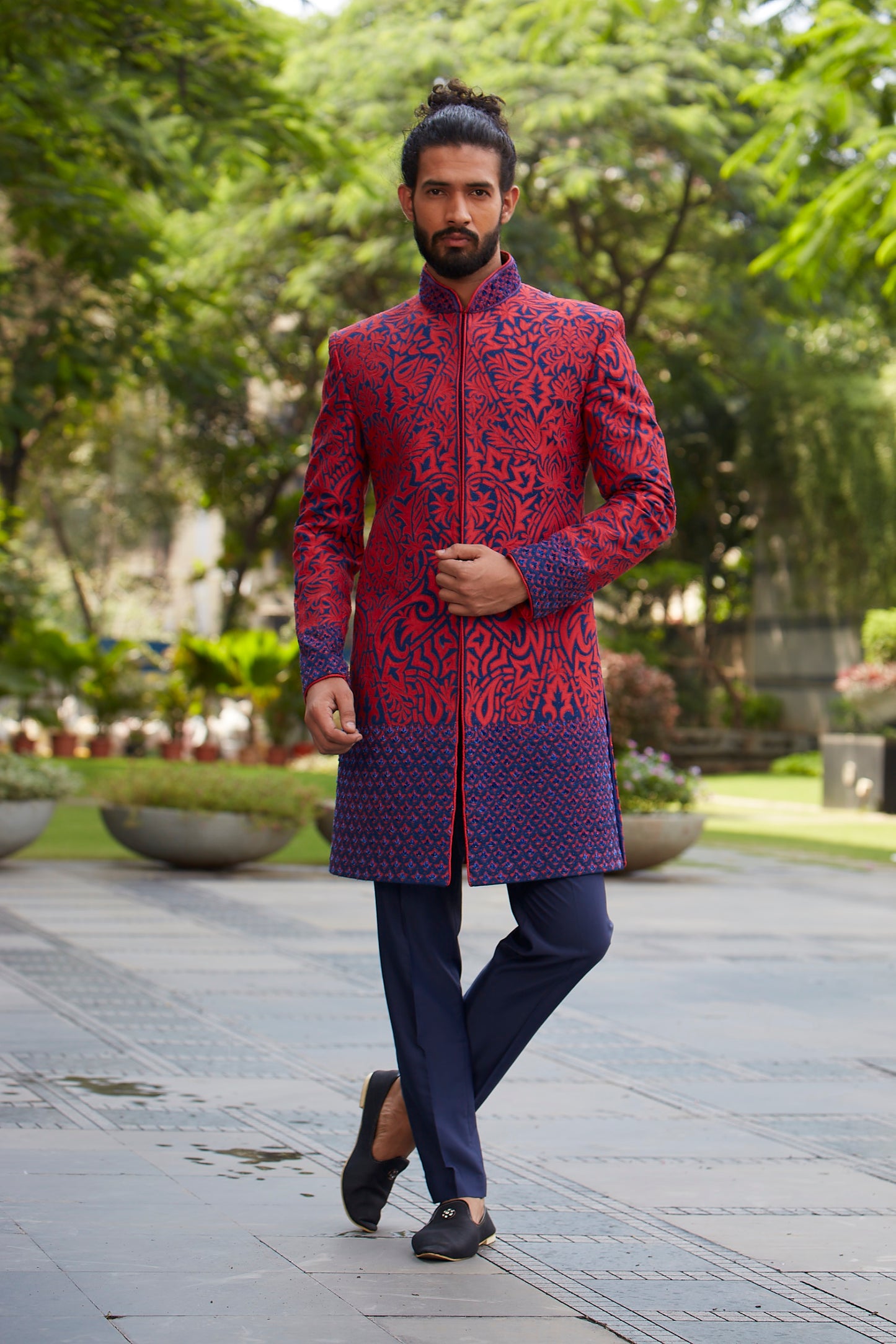 A man wearing a red and navy blue patterned sherwani jacket with navy trousers, walking on a paved path in a park with green trees in the background.