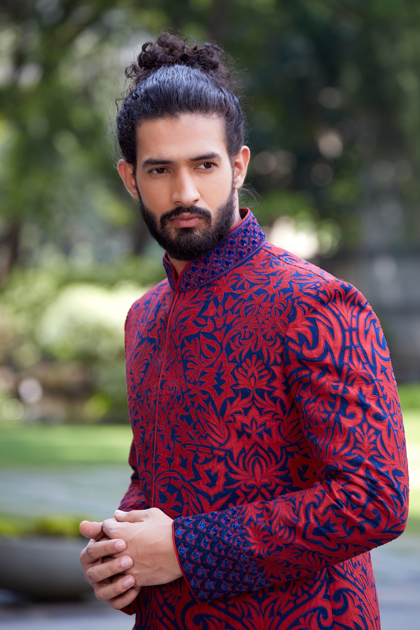 A man with a bun hairstyle and beard wearing a red and navy blue patterned sherwani jacket, standing outdoors with a blurred green garden background.