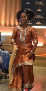 A woman modeling a traditional South Asian outfit in a metallic bronze-brown color with gold embroidery, standing on a runway.