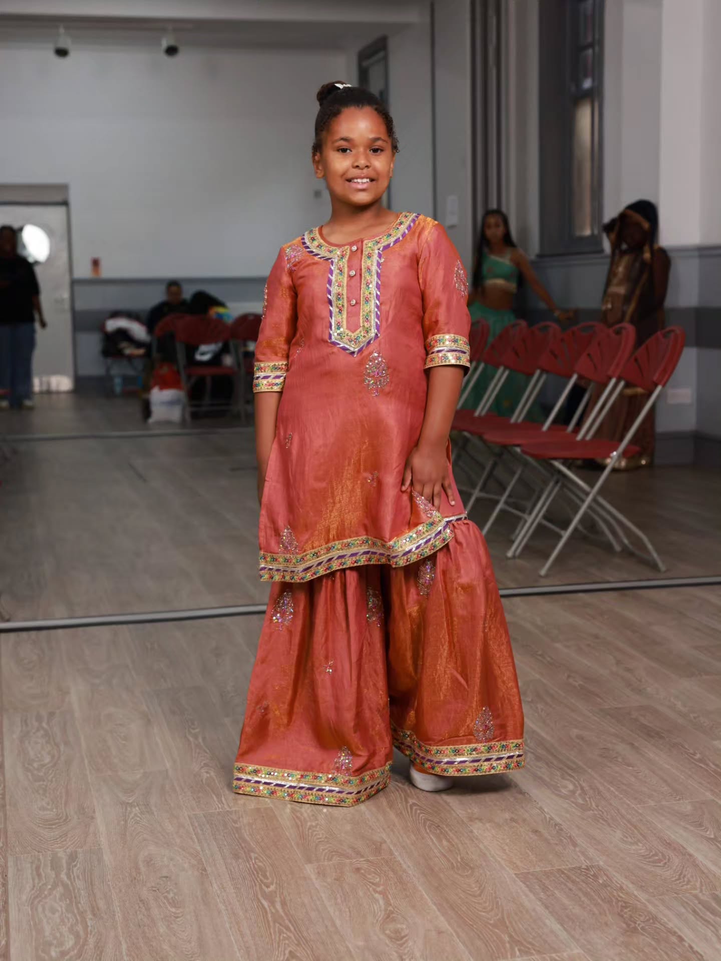 A young girl stands in a dance studio wearing a traditional Indian salwar kameez outfit in terracotta orange with gold and green embroidery. The background shows a mirror reflecting the room and other people.